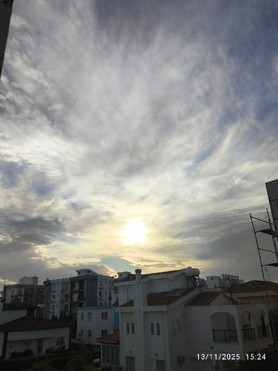 The image depicts an urban residential scene in Nicosia, Northern Cyprus / Cyprus, timestamped 13/11/2025 15:24. The sky occupies the upper two-thirds of the frame, characterized by a mix of cirrus and stratocumulus clouds, displaying hues of white, grey, and light blue. The sun is a bright, intense light source positioned in the upper-middle right, partially obscured by clouds. The lower third of the image shows a density of buildings. In the immediate right foreground, a multi-story white house with a red-tiled roof, multiple windows, balconies with dark railings, and a roof-mounted solar water heater system is prominent. Partially visible metal scaffolding abuts the far right edge of the frame. In the midground and background, a cluster of multi-story apartment buildings, primarily white and grey with flat roofs and balconies, are visible. Sparse green vegetation is interspersed among the structures at ground level. No individuals or discernible activities are present in the scene.