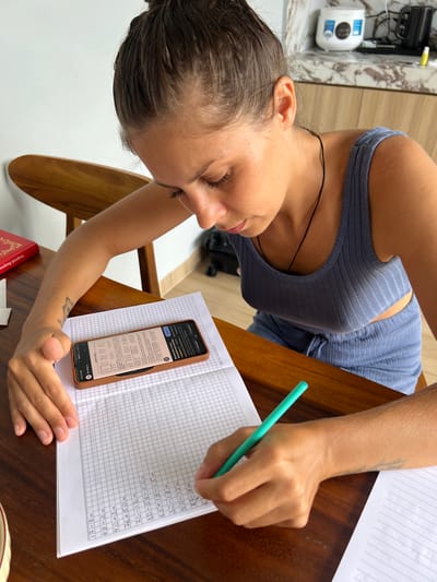 A woman sits at a wooden table, studying a math worksheet. The worksheet contains addition and subtraction problems. She is using a light green pencil to write on the paper. A smartphone displaying math problems from "My-shop.ru" is beside the worksheet. A book with gold lettering is visible near the bottom of the image. The woman is wearing a blue top. The background includes a light-colored wall and a white appliance with blue details. No information indicates this location to be Kutuh, Indonesia.
