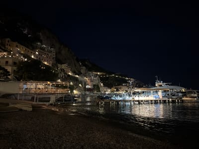 The image depicts a night scene in Pogerola, Italy. The foreground shows a rocky beach leading to calm, dark water. A pier extends into the water, illuminated with lights and lined with various boats, including a large yacht. Buildings and houses are built into the hillside, with lights visible in windows and along pathways. The sky is dark, with a few visible stars.