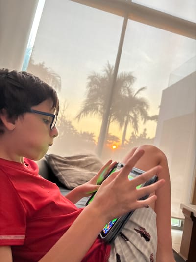 A boy with dark hair and glasses lies on a surface, dressed in a red shirt and striped pajamas. He is engrossed in a handheld gaming device, his hands actively engaged with the screen. Behind him, a window reveals the silhouettes of palm trees against a bright, sunset-like sky. The light filters through the window and reflects on the screen, creating a unique ambiance.