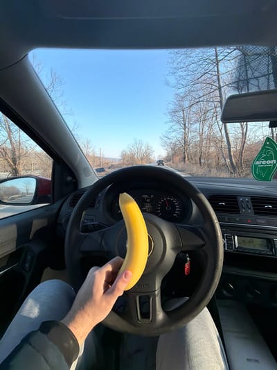 The scene depicts the interior of a moving vehicle, viewed from the driver's perspective, on a road in Komarevo, Bulgaria. A person's right hand, wearing a dark sleeve, holds an unpeeled yellow banana upright over the black steering wheel. The person's lap, covered by light gray sweatpants, is visible below the wheel. The car's instrument cluster shows a speedometer needle indicating approximately 50-60 km/h, with a digital display showing "20:06" and "25°C". A green "Areon" air freshener hangs from the rearview mirror. The car's radio system displays "N-JOY." Outside the vehicle, a clear blue sky extends above a two-lane asphalt road. Both sides of the road are lined with bare deciduous trees. A dark vehicle is visible in the distance on the road ahead, with distant terrain on the horizon. A dark rectangular object, possibly a sign or solar panel, is situated off the left side of the road.