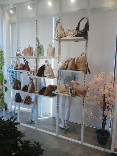 The image depicts a retail display of handbags in a store, likely a boutique or department store. The handbags are arranged on white shelving units, which are part of a modular display system. The shelves are tiered and have a clean, modern aesthetic.
The merchandise includes a variety of handbag styles, materials, and colors. There are tote bags, shoulder bags, and smaller clutch-like bags. Some bags are made of leather or faux leather, while others appear to be made of fabric or straw. Textures and embellishments vary, with some bags featuring intricate patterns, studs, or fringe. The color palette ranges from neutral tones like beige, tan, and white, to darker shades like brown and black, with some brighter accent colors present.
In addition to the handbags, the display includes a few mannequins dressed in light blue garments, likely to showcase how the bags might be styled. There are also decorative elements such as a potted artificial cherry blossom tree with pink flowers, and some greenery on the left side of the image. The overall ambiance suggests a well-organized and aesthetically pleasing retail environment.
The location is identified as Ávila, Spain, implying this is a store within that city. The background shows a white wall, and the lighting appears to be a mix of overhead track lighting and natural light filtering through blinds or sheer curtains. The floor is a neutral-toned tile.