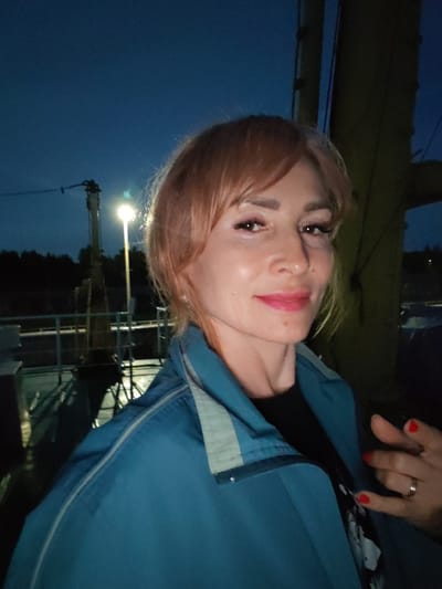 A woman with blonde hair and pink lipstick poses for a selfie at dusk in Sheksna, Russia. She wears a blue jacket over a black shirt. Her right hand, with red nail polish, is visible on the right side of the frame. In the background, a lamppost illuminates a dark sky and a railing along what appears to be a waterfront or dock.