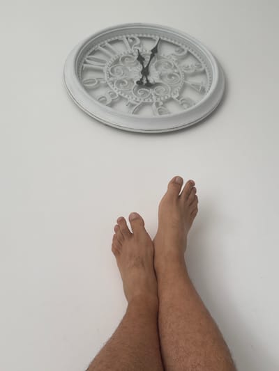 The image shows a close-up of a white decorative clock with Roman numerals on a white wall. The clock's hands indicate a time between 4:00 and 4:05. To the right of the clock, a pair of human legs and feet are visible, crossed at the ankles. The legs are hairy, suggesting they belong to a male. The feet are bare. The scene is indoors, with a plain white background and overhead lighting, creating a simple and uncluttered aesthetic. There is no visible text in the image. The composition and focus on the clock and feet suggest a possible theme related to time or waiting. The location context provided is Molenbeek-Saint-Jean - Sint-Jans-Molenbeek, Belgium, but this is not visually discernible from the image itself.