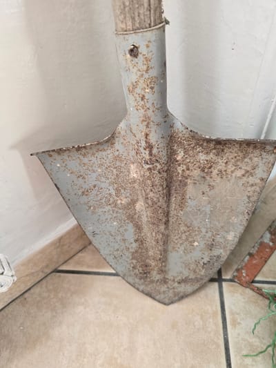 The image shows a close-up, waist-level, vertical shot of a rusty, grey metal shovel with a wooden handle. The shovel is positioned against a plain, off-white wall. The floor beneath the shovel is tiled with large, beige square tiles separated by dark grout lines. The shovel itself is heavily corroded, with significant patches of rust and discoloration. A circular emblem or marking is visible on the metal blade, though it is partially obscured by rust. To the right of the shovel, a portion of another object, possibly a piece of red-painted metal, is visible on the floor. A few strands of green, artificial-looking plant material are also present on the floor to the far right. The lighting is even, casting no distinct shadows. The scene appears to be indoors, possibly in a storage area or a less-trafficked part of a building. There are no people visible in the image, and no specific details suggest a location within the city of Salerno, Italy.