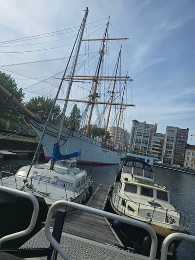 The image depicts a maritime scene in Ostend, Belgium. The foreground features a sailboat and motorboat docked beside a pier. In the background, a large three-masted sailing ship with intricate rigging stands prominently. Behind the ships, modern brick buildings line the waterfront. The sky above is partially cloudy.