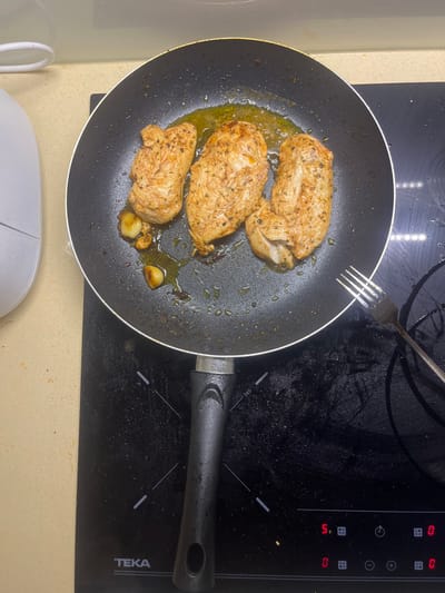 This image depicts a cooking scene within a kitchen located in Pedrouços, Portugal. The central subject is a black non-stick frying pan, positioned on an active black induction cooktop. Inside the pan, three pieces of seasoned chicken breast are cooking in a small amount of oil, alongside several cloves of garlic. The chicken appears partially browned and coated with visible herbs and spices.
The induction cooktop, manufactured by TEKA, displays red digital readouts, with "5." visible on the left burner control and "0" on the right. The cooktop surface shows some splatters of oil or liquid. To the right of the frying pan, a silver metal fork rests on the cooktop. To the left of the cooktop, a section of a light-colored, possibly beige or cream, countertop is visible. A portion of a white appliance, with a rounded, smooth exterior, is situated on the countertop in the upper-left quadrant of the frame. No individuals are present in the image.