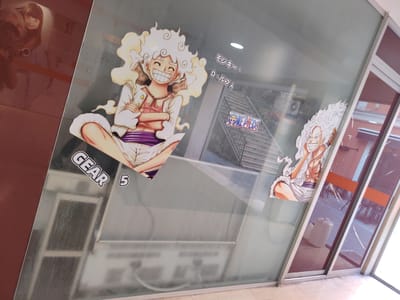 This image shows a street-level window display with anime-style stickers of characters from the series One Piece. Two large stickers of a character with white, curly hair and a smiling face are prominent, one on the left side of the window and a partial view of another on the right. The character on the left is wearing a white jacket over a pink shirt and purple shorts, with "GEAR 5" text below it. Japanese text, possibly the character's name "Monkey D. Luffy", is visible above the right sticker. A small "One Piece" logo is also present. The window reflects the interior of a building, revealing a staircase and what appears to be an air conditioning unit. The exterior view through the window shows a street with a doorway, a white plastic container, and tinted glass doors that reflect the environment outside. The overall scene suggests a shop or establishment in a city. Based on visual cues such as architectural style and the presence of a "Made in Italy" sticker visible in the original image, the location can be identified as Salerno, Italy.