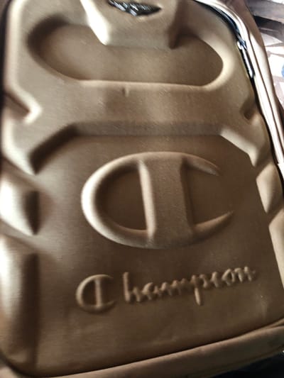 A close-up, rotated shot displays the textured, brownish-khaki or olive drab exterior of a bag or case. The material appears to be fabric, possibly a durable synthetic like nylon or polyester, exhibiting a subtle diagonal weave pattern. Along one edge, a metallic zipper is partially visible.
The surface of the bag features large, raised, embossed elements. Prominently displayed is the iconic "C" logo of the brand Champion, also embossed. Further along the bag, the word "Champion" is embossed vertically, with the letters "hampion" clearly discernible. The embossing creates distinct shadows, highlighting the three-dimensional nature of the branding.
The background is out of focus and indistinct, suggesting an indoor setting with no specific identifying features. There are no people or discernible activities taking place. The time of day and weather cannot be determined from the image.