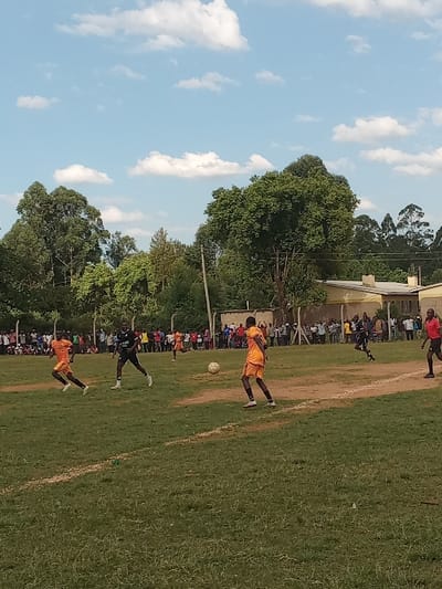 An outdoor football match is underway on a mixed grass and dirt field in Chevaywa ward, Kenya, beneath a light blue sky with scattered white cumulus clouds. Five male football players are visible. One player in an orange jersey and shorts runs from left to right in the foreground. Another player in a black jersey and shorts stands to his right, facing the center. A third player, also in an orange jersey and shorts, is positioned right of center, actively engaging with a light-colored football which is airborne near his feet. Two additional players, one in orange and one in red, are in motion further right and in the mid-background. A large crowd of spectators lines the background perimeter of the field, standing behind a fence. Beyond the spectators, dense green trees and light-colored buildings with dark roofs are present. A dark utility pole extends vertically on the right side of the scene.