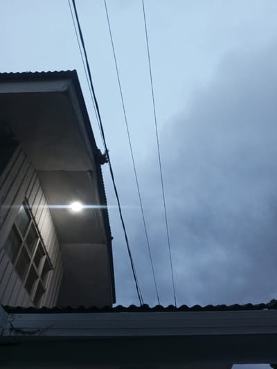 The scene, situated in B. Nam Ngao, Laos, presents an upward-angled exterior view under an overcast, blue-grey sky, suggesting dusk or dawn. A section of a light-colored building with vertical panel siding and a multi-paned window is visible in the upper left quadrant. A single, bright white artificial light source, likely a bulb or LED, is affixed to the underside of the building's eave, projecting a distinct lens flare. Above this light and extending rightward, a dark corrugated metal roofline is visible. Multiple black utility lines, appearing to be power or communication cables, traverse diagonally across the sky from the left to the mid-right. In the lower central portion of the frame, a second, lower dark corrugated roofline with an attached light-colored fascia or gutter system is present. No individuals are depicted in the image.