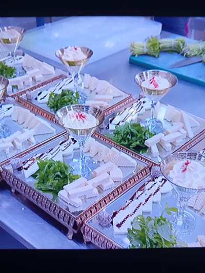 The image shows a close-up, angled view of several mirrored trays filled with cheese platters. The platters contain various types of cheese, some cut into rectangular blocks and others appearing as more solid pieces. There are also small bunches of green leafy herbs, likely for garnish, placed on the cheese. Some of the cheese is drizzled with a dark sauce, possibly balsamic glaze, creating a decorative pattern. In the background, out of focus, are several martini glasses filled with a white, creamy substance topped with a red garnish. To the right of the platters, a bunch of celery stalks tied with white twine is visible on a blue cutting board, along with a knife. The overall setting appears to be a kitchen or preparation area, with a stainless steel countertop. The image is a still from a video, indicated by the pixelation and slight blurriness. There are no people visible in the frame. The location in Pavlohrad, Ukraine is not discernible from the visual information provided in the image.