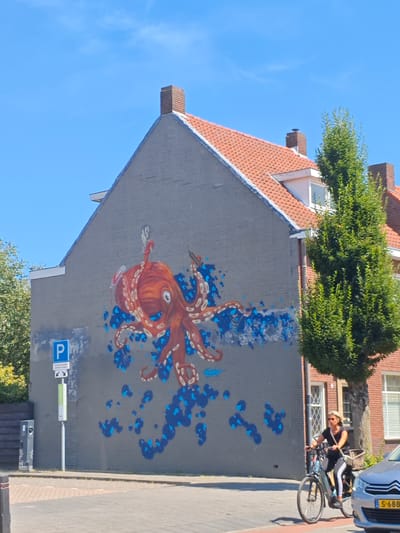 An outdoor, eye-level shot shows a grey brick building with an orange octopus mural painted on its side. A woman wearing sunglasses, a black top, and striped leggings is riding a bicycle with a basket on the front. A silver car is parked to the right of the woman. A tree with green leaves is positioned to the right of the building. A blue and white parking sign is visible on the left side of the building. The sky is clear and blue. The image appears to be taken in Tilburg, Netherlands.