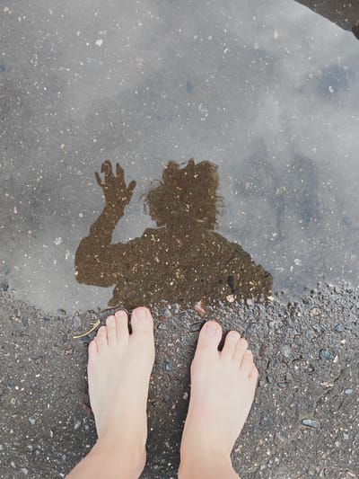 A reflection of a person is visible in a puddle on an asphalt surface, with the person's bare feet in the foreground. The reflection shows the person's head and raised right hand. The puddle is on a dark, wet asphalt ground with small stones and debris scattered on it. The scene appears to be in a public area, possibly a street or sidewalk, and the lighting suggests it is daytime. The location is identified as Koledino, Russia.