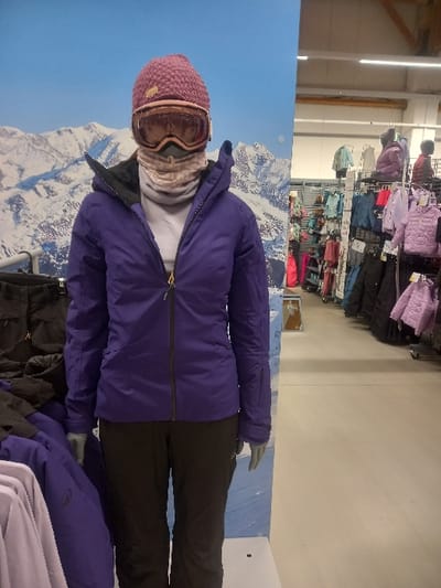 An upright image displays a mannequin showcasing winter sports attire within a retail store in Vendin-le-Vieil, France. The mannequin is dressed in a dark purple insulated winter jacket with a black zipper, and black winter pants. It wears a textured pink or mauve knit beanie hat, over which a pair of light-framed ski goggles with tinted lenses are positioned. A light-colored neck gaiter covers the mannequin's lower face and neck, with a white or light grey base layer visible beneath the open jacket.
Behind the mannequin, a large backdrop panel features a scenic image of snow-capped mountains under a clear blue sky, simulating a winter alpine environment. To the right, the interior of the store reveals multiple retail racks filled with various colorful winter jackets and clothing items, predominantly in shades of purple, pink, light blue, and green. The store has a white ceiling with fluorescent lighting and light-colored walls. A portion of another mannequin, dressed in a similar purple jacket and black pants, is visible in the bottom right corner, indicating a larger display area for winter apparel. The scene captures a retail setting dedicated to winter sports clothing and equipment. No discernible text is visible in the image.
