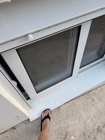 A person's left foot, wearing a navy blue flip-flop with "TOMMY HILFIGER" written on the strap, is positioned on a white windowsill. The foot is stepping onto the windowsill, which appears to be part of an open window. The window frame is white and the glass is tinted dark. Below the open section of the window, there is a dark interior. The windowsill is situated next to a textured, light-colored concrete surface. The scene suggests an outdoor setting, possibly during the daytime. The image has a slightly washed-out appearance, possibly due to bright sunlight. The backdrop consists of a white wall and a rough concrete area. The overall impression is that of a casual, everyday moment.