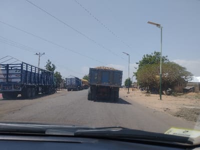 The image shows a road in Bajoga, Nigeria (latitude: 10.91, longitude: 11.472).  There are three large trucks traveling on the paved road, one of them loaded with what appears to be large sacks of grain.  The truck in the foreground is partially obscured by the windshield of a car, and a few other vehicles can be seen further down the road.  There are two street lights on the right side of the image, and a few trees with sparse leaves are on the right side of the road.  The road is bordered by dirt shoulders, and there are a few low-lying buildings in the distance on the right. The image was taken on a clear day with a blue sky.