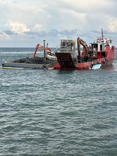 The image captures a maritime material handling operation offshore in La Ceiba, Honduras. In the foreground, choppy, dark grey-green water is visible under an overcast sky with extensive grey cloud cover.
Two primary vessels are featured:
1. **Red Vessel (Right):** A large, red-painted vessel, possibly a landing craft or self-propelled barge, displaying significant wear and rust. Its bow ramp is lowered towards the water. A yellow excavator, identifiable as a Caterpillar by its distinct coloration and design, is positioned on the vessel's deck, facing forward with its boom extended. Several individuals are present on board; one person in an orange safety vest stands near the lowered ramp, alongside a small, light blue and white fiberglass dinghy moored to the vessel's starboard side. Additional crew members are discernible near the stern's pilothouse structure.
2. **Grey Barge (Left):** A flat-decked, grey barge is situated adjacent to the red vessel. It is heavily loaded with a substantial pile of dark, granular material, consistent with dredged sediment or aggregate. Yellow perimeter fenders are visible along its sides. An orange excavator, bearing partial "Link-B..." text (likely indicating a Link-Belt model), is positioned on the grey barge, with its boom extended over the material pile. The barge appears to be riding low in the water due to its cargo.
The close proximity and configuration of the two vessels, along with the excavators and material, indicate an active process of dredging, loading, or transferring bulk material between them.