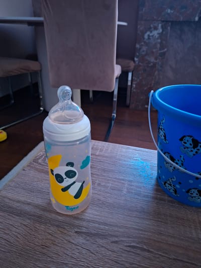 A baby bottle with a panda graphic and the "Tigex" logo is placed on a wooden table. Next to the bottle is a blue bucket adorned with Dalmatian dog illustrations. In the background, chairs are visible. The scene is indoors.