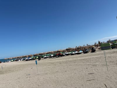 The image depicts a beach scene in Năvodari, Romania, on a clear, sunny day. The foreground is dominated by a sandy beach with tire tracks indicating vehicle activity. Two green poles with plastic bottles attached are planted in the sand. In the mid-ground, a line of jet skis is parked, some on trailers. Behind the jet skis, a row of thatched umbrellas provides shade, with lounge chairs and what appear to be stacked mattresses visible beneath them. In the background, the sea is visible, and further in the distance, buildings and palm trees suggest a resort area. The sky is a vibrant, cloudless blue. There are no people visible in the immediate vicinity of the jet skis and umbrellas.