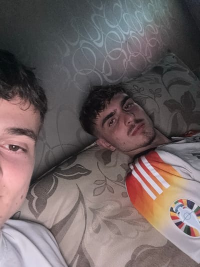 The image depicts two young adult males in an indoor setting, likely on a couch or bed, under dim lighting conditions.

One male is partially visible on the left, showing his left eye, eyebrow, and a portion of his face with dark brown curly hair and fair skin. He is positioned close to the camera, with his eye directed towards it, suggesting he is holding the device used to capture the image. His attire consists of a plain light-colored, possibly white, t-shirt, with only the shoulder and upper arm visible.

The second male is centrally positioned, lying on his back with his head resting on a patterned pillow. He has dark brown wavy hair, brown eyes, and light stubble on his chin and upper lip. He is wearing a light-colored t-shirt, possibly white or light grey, which features a distinct graphic on the right shoulder/sleeve area. This graphic includes several vertical stripes in shades of red, orange, and yellow, blending into one another, alongside a circular emblem. The emblem consists of a stylized trophy-like cup centered within a target-like design, surrounded by a rainbow of colored segments, identifiable as the UEFA Euro 2020 tournament logo. His gaze is generally directed towards the camera.

The background consists of patterned upholstery or fabric. Directly behind the second male's head is a cushion or pillow with a light beige base featuring dark brown/grey stylized leaf and vine patterns. To the upper left, a darker, possibly wall-mounted or upholstered surface displays a lighter, repeating motif of abstract, interconnected swirling lines. The ambient light is subdued, creating soft shadows and highlights on the subjects' faces and surrounding textures. No discernible features within the image indicate a specific city or country.