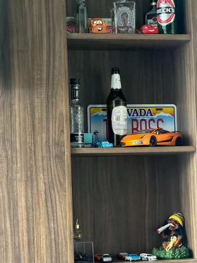 The image shows a close-up of a wooden shelf filled with various items. The shelf is divided into compartments. In the left compartment, there are several bottles and small figurines. One bottle is labeled "BECK'S" and another appears to be "BACAR." There is also a small figurine of the character Lightning McQueen from the movie "Cars." Another item in this section is a bottle with a silver horseshoe emblem and the text "HERRADURA ANEJO."

The middle compartment contains more bottles and a license plate. One bottle is labeled "Bitburg Premium A" and is from Germany. Behind the bottle is a license plate with the text "VADA ROCC" and a mountainous landscape. A bright orange toy car is placed on top of the license plate. There is also a small blue toy car. Further down, there are more bottles, one with the label "MAESTRO DOBEL DIAMANTE."

The right compartment of the shelf features a small figurine of a person smoking, with yellow and red headwear. There are also several miniature toy cars lined up vertically. The background of the shelf is dark wood. The overall scene appears to be an indoor collection of items, possibly a display on a shelf in a home or a bar. The lighting suggests an indoor environment, possibly in the evening or with artificial light.