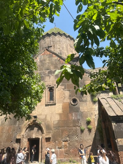 In Bjni, Armenia, a group of people stand before the ancient stone facade of a church, partially obscured by lush green tree branches against a clear blue sky. The church, constructed from rough-hewn reddish-brown stones, features a prominent arched entryway with a dark opening. Above the entrance, the weathered stone walls are adorned with inscriptions, and a small, rectangular window is visible to the left. A cylindrical opening is set into the wall to the right of the window, and a pointed structure with a cross on top, covered in grass, rises above the main structure. Some individuals are entering or exiting the church, while others observe its exterior. A young girl in a yellow shirt stands near a man wearing a light-colored cap and a white t-shirt. The scene is bathed in bright sunlight, casting shadows on the stone.
