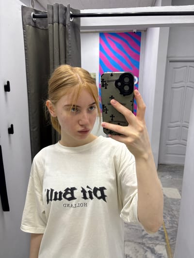 A young woman with blonde hair and light skin is taking a selfie in a brightly lit room. She is wearing a white t-shirt with a black gothic-style font graphic on it, which appears to read "DIRT BUM" upside down and "MUD BUM" right-side up. She is holding a smartphone in her right hand, which has a camera case with a pattern of crosses. Her hair is styled in loose waves, and she has a neutral expression on her face.
The background of the image is somewhat blurred but shows a wall with a large, colorful graphic featuring pink and blue wavy lines. To the left, a white column and a grey curtain are visible. The floor appears to be white. The room seems to be indoors, with diffused lighting suggesting it could be daytime. The overall impression is casual and personal, typical of a selfie taken in a home environment. No specific weather conditions are discernible. The location context provided is Kuznetsk, Russia.