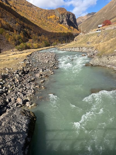 The scene is a mountainous river valley in Kvemo Okrokana, Georgia, observed under a partly cloudy sky. A light green, turbulent river, exhibiting rapids and white foam, flows from background to foreground. The river's left bank features a stony riverbed and an ascending slope densely covered with trees displaying yellow and orange autumn foliage. The right bank, characterized by dry grass and scattered rocks, ascends steeply. High on the right slope, a white sport utility vehicle is parked on a dirt track adjacent to a small structure with a dark roof. A tree with red foliage is positioned nearby. Additional metallic structures are present lower on the right slope. A large, textured dark grey rock occupies the lower left foreground, casting a shadow partially over the water. No individuals are visible.
