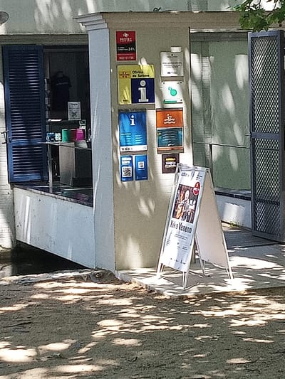 The image shows the exterior of a building with several signs. The building appears to be an information point or a small shop. In the foreground, an A-frame sign displays a poster with the text "Kiko Veneno" and a photograph of a man playing a guitar. Behind this sign, the building has a blue shuttered window to the left and a metal-grated door to the right.

On the wall of the building, there are several smaller signs. One red sign has a logo and text that reads "PROTEC 24h". Below this, a yellow sign reads "Oficina de Turismo" (Tourist Office). Further down, there are blue signs with an "i" icon, indicating information. Other signs are visible but illegible due to image quality.

Inside the building, through the open window, a person wearing a dark t-shirt can be seen. Colorful containers, possibly cups or lids, are visible on a counter. The ground in front of the building is sandy and covered in dappled sunlight and shadows, suggesting an outdoor area. The architecture and signs suggest this is a public service building located in Banyoles, Spain, likely a tourist information center.