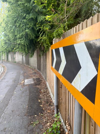 The image depicts a street scene in Guildford, United Kingdom. A section of roadway is visible, featuring a black asphalt surface. A triangular road sign with reflective white and black directional arrows, bordered in orange, is affixed to a wooden structure. A section of the wooden structure is above a concrete wall, with shrubbery and trees bordering the other side. The scene appears to be in an outdoor environment with natural lighting. No people are visible in the image.
