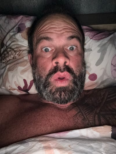 A man with a graying beard and furrowed brow lies on a floral-patterned pillow beneath a patterned duvet. The man's mouth is pursed. A tattoo is visible on the man's chest. The image seems to have been taken indoors and the location cannot be determined from the image. The photograph does not show the city of Marche-en-Famenne, Belgium.