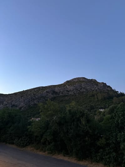 An upward angle shot captures a steep, rocky mountain face covered in dense, dark green foliage. The mountain dominates the mid-ground and extends to the right of the frame. A clear, pale blue sky fills the upper portion of the image. In the lower right quadrant of the mountain, a white building with a reddish-brown roof is partially visible through the trees. A dark, paved road with a gravel shoulder runs along the bottom edge of the frame, indicating a road leading away from the mountain. The scene appears to be set in Anduze, France, likely during dusk or early dawn given the soft light and clear sky. There are no people or animals present in the image.