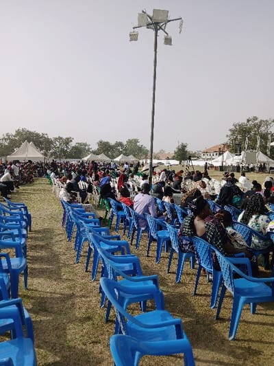 Rows of blue plastic chairs are filled with people outdoors on a grassy field. A tall metal pole with multiple floodlights stands prominently. Tents are visible in the background among the trees. The crowd extends into the distance. The sky is pale and overcast.
