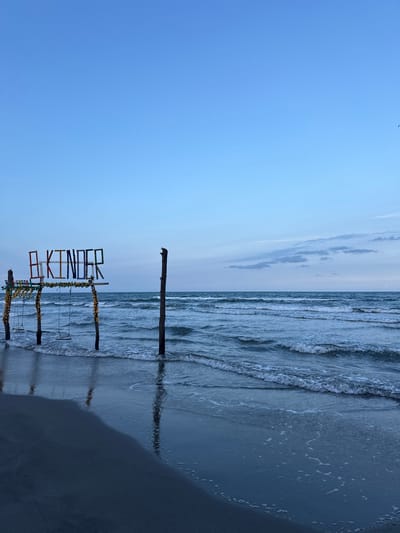 The image shows a beach scene, likely in Mata de Uva, Mexico. A wooden structure with the word "KINDER" in colorful lettering stands on the shore. The structure has swings hanging from it. The beach slopes into the ocean, with waves visible. Two weathered wooden poles stand further back, also partially submerged in the water. The sky is light blue, with some clouds. The overall lighting suggests it's daytime, possibly morning or late afternoon.