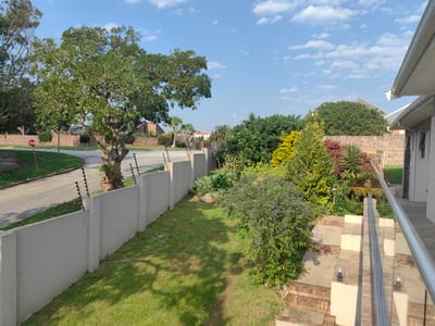 This is a photograph of a suburban residential area in Port Elizabeth, South Africa, likely taken in the late afternoon given the long shadows cast by the sun. The scene depicts a well-maintained property with a focus on the garden and its surrounding features.
In the foreground, a light-colored concrete wall with an electric fence atop runs along the property line, separating the yard from a paved road. A large, leafy tree with a thick trunk stands near the road, and a red "STOP" sign is visible on a pole to its left.
The yard itself is covered in green grass, with a path leading through it. A mature bush with small purple flowers is prominent in the middle of the yard. Beyond the wall, more lush vegetation and trees are visible, contributing to a sense of verdant landscaping.
To the right of the frame, a modern house with a white exterior and a white roof overhang is partially visible. A set of outdoor stairs, constructed with a combination of brick and light-colored stone, leads up towards the house. A glass and metal railing lines the stairs and a balcony area.
The sky is a clear blue with scattered white clouds, suggesting pleasant weather. The overall impression is one of a tranquil and secure residential environment. There is no visible activity of people taking place, and no discernible emotions are conveyed. The visible text is limited to the "STOP" sign.