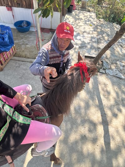 A man in a red "Việt Nam" hat is shown in an eye-level shot, standing beside a horse with a red head ornament. The horse is saddled, and someone wearing pink leggings and black clothing is sitting on the horse. The location is likely in Bắc Hà, Vietnam, though it is not definitively shown. The setting appears to be an outdoor area with concrete paving and some vegetation.