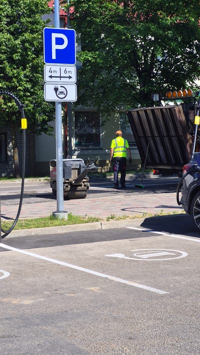 A street cleaner wearing a yellow high-visibility vest and an orange hard hat is sweeping the sidewalk with a broom. The street cleaner is standing next to a parked construction vehicle. In the background, a garbage truck is visible, along with trees and buildings. Several road signs are also present, including a blue parking sign with the letter "P" and a white sign with an electrical plug symbol. The image appears to be taken during the daytime, under clear weather conditions. The scene is set on a street in Līvāni, Latvia, indicated by the presence of Latvian text on a sign, "MANA APTIEKA," which translates to "My Pharmacy."
