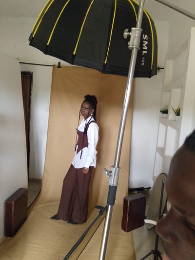 This is a medium shot photo taken indoors, likely in a photography studio, with a neutral brown backdrop. The subject is a young woman of African descent, standing and posing for a photo. She has her hair styled in intricate braids, with some strands falling forward. She is wearing a white collared shirt with a dark brown corset or vest over it, and matching wide-leg brown pants. She also appears to be wearing dark shoes. Her stance is confident, with her left leg slightly forward and her body angled towards the viewer. She is wearing earrings and a watch on her left wrist.
In the background, a large softbox light with yellow accents is positioned overhead and to the left of the subject, indicating a professional lighting setup. A silver lighting stand is prominently in the foreground, partially obscuring the subject. To the right of the subject, another stand with a reflector is visible, along with a dark rectangular object and a blurred out head in the foreground, suggesting the presence of a photographer or other crew. The walls of the room are white, with some shelves visible on the right wall holding small potted plants. The floor appears to be a light wood or linoleum. The overall impression is that of a planned photoshoot. The visible text on the softbox reads "SML 105". The time of day and weather are not discernible from the image. The location context is provided as Anguldi, Nigeria.