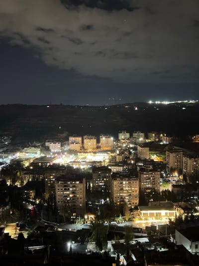 An aerial view at night reveals a city nestled along a dark coastline beneath a cloudy sky. Bright city lights illuminate numerous buildings, streets, and a few trees. The skyline features densely packed structures, indicating a thriving urban environment. The contrast between the dark sea, the mountains, and the bright city creates a captivating scene.