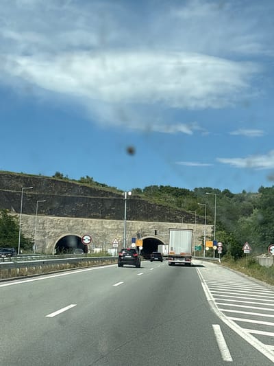 The image captures a view from a vehicle traveling on a highway towards a tunnel entrance. The tunnel is carved into a hillside covered with dark, rocky material and vegetation. The highway is paved with asphalt and has clear lane markings. Several vehicles are visible on the highway, including a white truck, a black car, and other vehicles in the distance. Traffic signs, including speed limits and tunnel entrance warnings, are posted along the side of the road. The sky is blue with scattered white clouds. The scene appears to be in Blatino, Bulgaria, given the context.