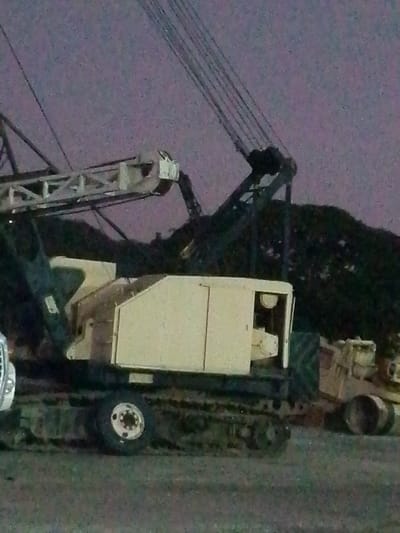 The image depicts a large, industrial crane situated outdoors, possibly in the Parroquia Guatire, Venezuela. The crane is primarily beige and black, featuring a tracked base, a large cabin, and a towering boom with visible cables. Another piece of heavy equipment, possibly a bulldozer, is visible to the right of the crane. The background appears dark, suggesting the image was taken at night or in low-light conditions. Behind the crane and bulldozer, there is a large, dark, undefined mass which could be a pile of material or vegetation. The ground is flat and appears to be paved or covered in dirt.