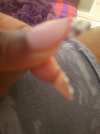 The image displays an extremely out-of-focus, close-up perspective of a human hand in Mumbuni North ward, Kenya. The visible elements consist primarily of a thumb and the distal phalanx of an adjacent finger, exhibiting a medium-brown skin tone. The thumbnail features a light pink or natural-toned artificial nail extension with a pointed or oval tip. The tip of the adjacent finger is covered with an opaque white substance. The hand is positioned over a grey fabric surface, possibly clothing, which shows visible folds. In the blurred background, a mass of crumpled dark purple fabric is visible next to a section of red and purple plaid fabric. A small, unidentifiable, light-colored object with dark elements is discernible but highly blurred near the tip of the thumb. No specific actions or interactions beyond the visible state of the hand and objects can be definitively ascertained.