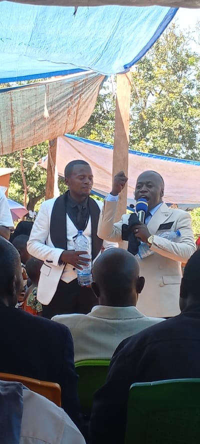 Outdoors in Kalemie, Democratic Republic of the Congo, two men in suits stand before a seated audience. The man on the left wears a white suit with black lapels and holds a water bottle. The man on the right, wearing a light-colored suit, speaks into a blue microphone and holds a black cloth and another water bottle. An overhead canopy provides shade. The audience consists of people in various attire, seated in green chairs. Trees are visible in the background.