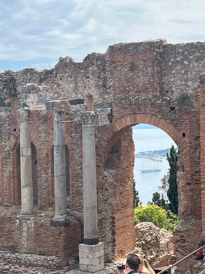 The image shows a view through an ancient Roman amphitheater in Taormina, Italy, overlooking a body of water with a large sailing ship. The amphitheater is constructed of brick and stone, with visible columns and arched doorways. The sky is partly cloudy. In the foreground, two individuals are present, with one appearing to hold a camera. The overall scene conveys a sense of historical significance and picturesque scenery.