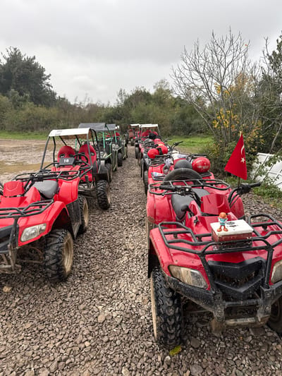 The scene is an outdoor staging area for off-road vehicles in Çiftlikköy, Turkey, under an overcast sky. A gravel path serves as the parking surface for multiple ATVs and UTVs/buggies, arranged in two irregular lines. The majority of the vehicles are red, with a few green UTVs visible in the background of the left line.
In the foreground, a red ATV on the right displays a small Turkish flag attached to its handlebar and a small, orange-capped figurine standing on a white box secured to its front cargo rack. Several red helmets are placed on the seats or cargo racks of the ATVs in this line, indicating preparation for or completion of a recreational activity. Another red ATV in the left foreground, behind the first, exhibits the number "16" on its front panel. All vehicles show evidence of prior use, with mud and dirt adhering to their tires and lower chassis.
The environment comprises dense, varied foliage and trees, some with bare branches or yellowing leaves, lining the sides of the gravel path. In the far right background, partially obscured by the trees, some discarded white panels or objects are visible on the ground. No individuals are present in the image.