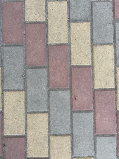 This is a top-down, close-up shot of a paved surface in Azov, Russia. The pavement is made of rectangular interlocking stones in three colors: red, yellow, and gray. The stones are laid in a running bond pattern, with each row offset from the one below. The surface appears to be outdoors and is well-lit, suggesting it is daytime. There are a few small pieces of debris, such as twigs and dirt, scattered on the stones, indicating it may be a public walkway or plaza. No people or specific landmarks are visible. The image focuses solely on the texture and pattern of the paving stones.