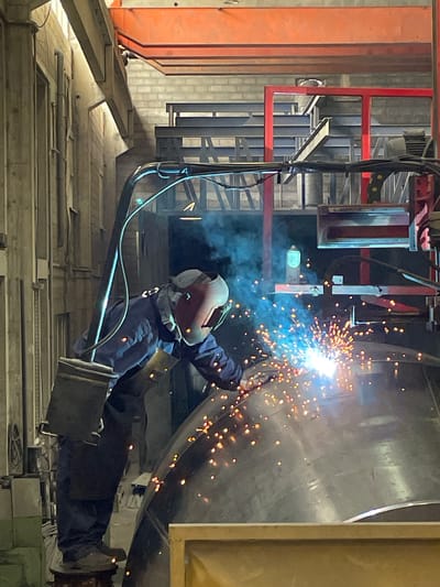 In Zaljevo, Montenegro, a welder in a blue uniform and protective mask is actively welding a large metal object in an industrial setting. The welder stands on a circular platform, holding a welding torch that emits bright sparks and blue smoke. The welding process is illuminated by the intense light from the torch, creating a fiery display on the metal surface. In the background, industrial machinery, including metal beams and structures, are visible, suggesting a manufacturing or repair facility. The environment is dimly lit, with overhead lighting casting shadows and highlighting the illuminated welding activity.