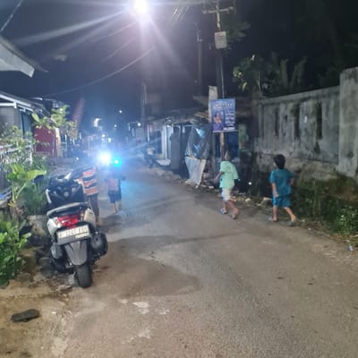 The image shows a street in the Kecamatan Tamansari neighborhood of Jakarta, Indonesia. It is nighttime, and the street is dimly lit. There are four people in the image: two children playing, one adult male standing near a motorcycle, and one unidentified person in the background. The children are running down the street, and the adult male appears to be watching them. The motorcycle is parked on the side of the street. The scene is also characterized by the presence of a high wall on the right-hand side of the street and a sign on a pole. There are plants and trees along the wall. The street is paved with asphalt and the ground is covered in some dirt and small plants. The image was taken from the perspective of someone standing on the side of the street, facing the direction of the people.  The street is relatively empty, except for the people and the motorcycle. The scene is quiet and peaceful, with the only sound being the sound of the children playing.