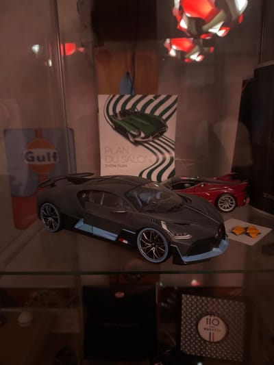 A glass shelf displays car-themed collectibles. A dark gray Bugatti model dominates the foreground. Behind it are a red car, a brochure titled "Plan du Salon," and a Gulf Oil emblem. To the right, stacked boxes feature Bugatti branding. In the background, a multi-colored, geometric lamp adds a warm glow. The arrangement suggests a hobbyist's collection showcasing their passion for automobiles, particularly Bugatti.
